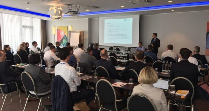 Eine Gruppe von Menschen sitzt in einem Konferenzraum und blickt aufmerksam auf einen Vortragenden, der neben einem Bildschirm steht, auf dem eine Präsentation zum Thema virtuelle Zusammenarbeit läuft. Der Raum ist modern eingerichtet und hat Deckenbeleuchtung, und die Teilnehmer hören aufmerksam zu.