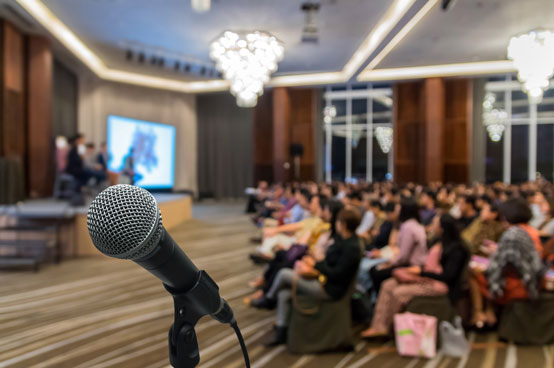 Ein Mikrofon im Vordergrund richtet den Blick auf einen Konferenzraum voller sitzender Teilnehmer. Im Hintergrund ist unter eleganten Kronleuchtern eine Gruppe von Rednern auf der Bühne vor einem großen Bildschirm zu sehen, der über MS Stream streamt.