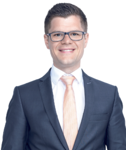 Ein Mann in dunklem Anzug mit heller Krawatte und Brille lächelt in die Kamera und strahlt Selbstvertrauen aus, als würde er sein Team mit Leichtigkeit führen. Der Hintergrund ist schlicht weiß und spiegelt eine Konzentration wider, die einer effektiven virtuellen Zusammenarbeit ähnelt.