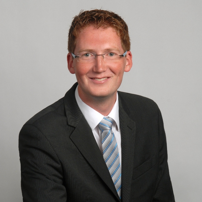 Eine Person mit kurzen roten Haaren und Brille lächelt, während sie einen schwarzen Anzug, ein weißes Hemd und eine gestreifte Krawatte trägt und den professionellen Geist der Leitung eines Teams verkörpert. Der Hintergrund ist in einer schlichten, hellen Farbe gehalten.