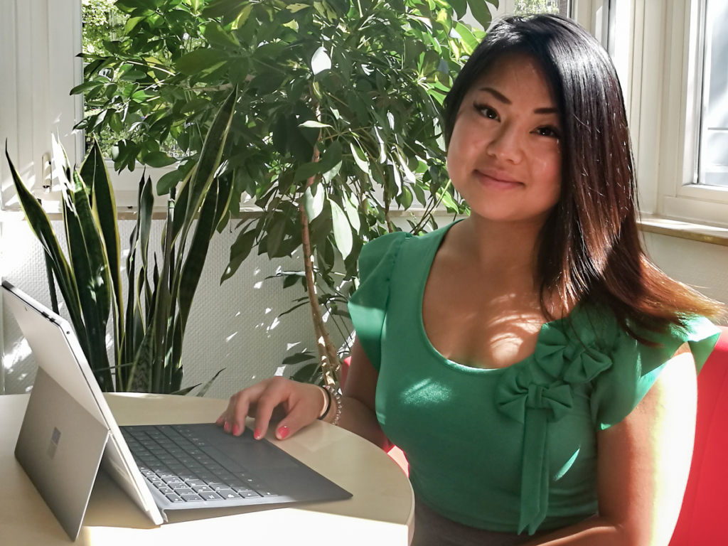 Eine Frau in einem grünen Oberteil sitzt an einem Tisch und nutzt ihren Laptop, um möglicherweise Microsoft Power BI zu erkunden. Sie lächelt in die Kamera, im Hintergrund sind üppige grüne Pflanzen und ein sonnenbeschienenes Fenster zu sehen.