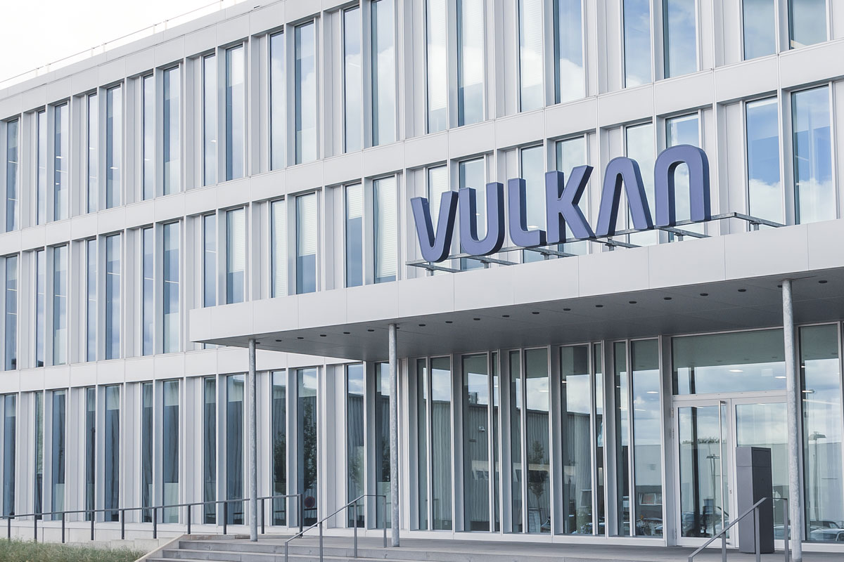Modernes Bürogebäude mit großen Glasfenstern und „VULKAN“-Schild über dem Eingang. Die Fassade zeichnet sich durch ein klares, minimalistisches Design mit vertikalen Linien aus, das an die rationalisierte Effizienz von Softwarelösungen erinnert. Stufen führen unter einem teilweise bewölkten Himmel zu den Haupttüren.