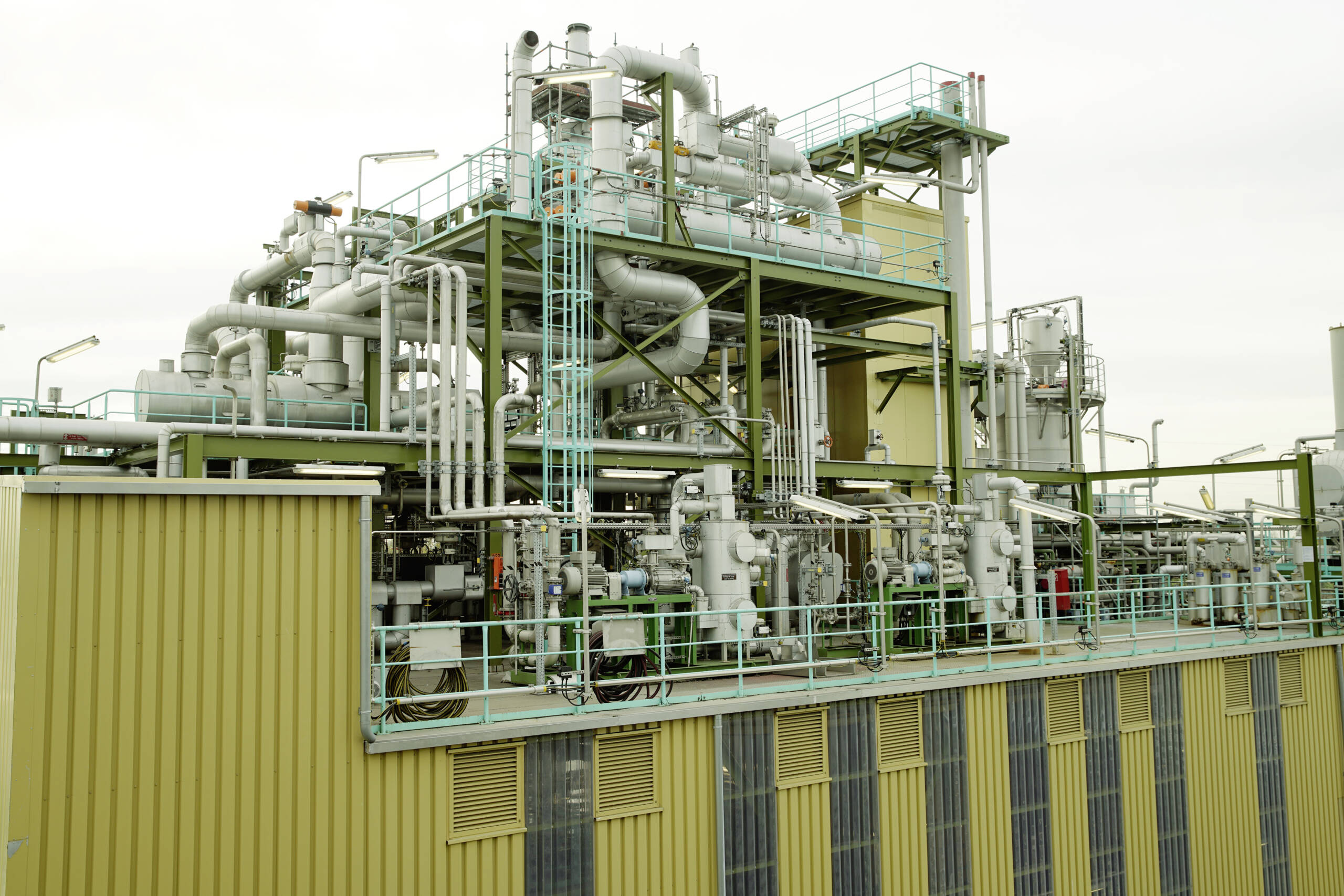 Eine Industrieanlage mit komplexen Rohrleitungen und Maschinen auf einem gelben Gebäude zeigt ihre komplexen Systeme. Die an Softwarelösungen erinnernde Struktur umfasst verschiedene Metallrohre, Ventile und Plattformen. Ein bewölkter Himmel bietet einen neutralen Hintergrund für die metallische Ausrüstung.