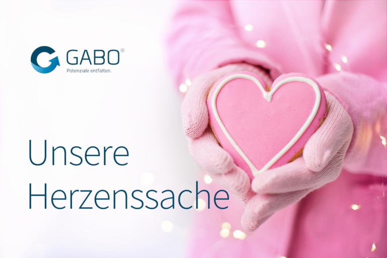 Eine Person in rosa Handschuhen und Mantel hält einen herzförmigen rosa Keks. Der Text lautet: "GABO Unsere Herzenssache". Weiße Lichter verleihen dem Bild eine festliche Note und weisen auf die Wärme der Microsoft-Intranet-Verbindungen hin.