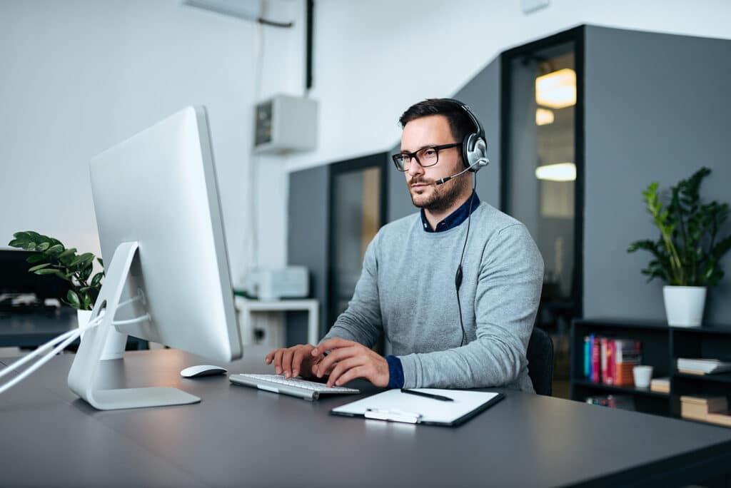 Ein Mann mit Brille und Headset arbeitet an einem Desktop-Computer in einem modernen Büro, möglicherweise in einer Microsoft-Schulung. Auf dem Schreibtisch befinden sich ein Klemmbrett, ein Stift und eine Pflanze. Im Hintergrund sind Regale und große Fenster zu sehen.