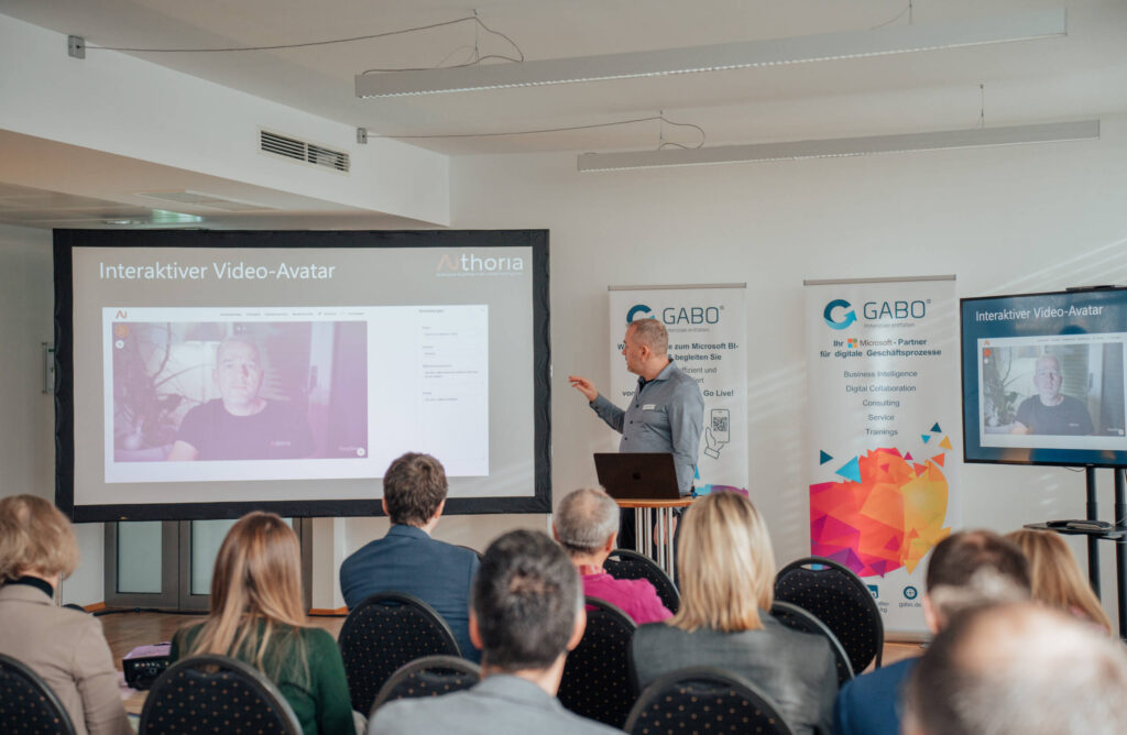 Ein Mann hält eine Präsentation vor einem Publikum in einem Konferenzraum und zeigt dabei auf einen Bildschirm, der eine in Microsoft 365 integrierte Video-Avatar-Schnittstelle anzeigt. Im Hintergrund sind zwei Banner mit Firmenlogos zu sehen.