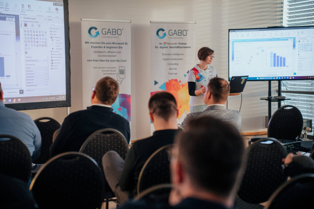 Eine Frau präsentiert den Zuhörern in einem Konferenzraum auf einem großen Bildschirm Diagramme, die Erkenntnisse aus dem Bereich Business Intelligence aufzeigen. Die Teilnehmer sehen sie an, während sie neben Bannern mit dem GABO-Logo und einem Laptop steht. Durch die Jalousien fällt natürliches Licht ein.