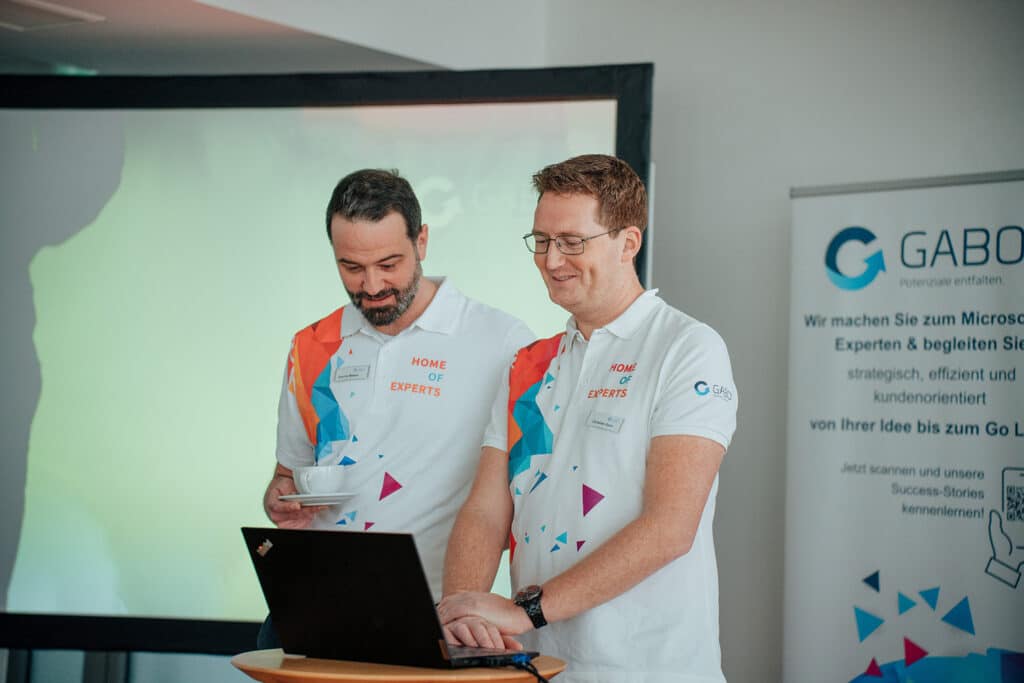 Zwei Männer in weißen "Home of Experts"-Shirts stehen an einem Laptop, einer hält eine Tasse mit Untertasse. Hinter ihnen sind ein GABO-Banner und ein Projektor zu sehen, während sie sich auf eine geschäftliche oder technische Diskussion über Microsoft Power BI konzentrieren.