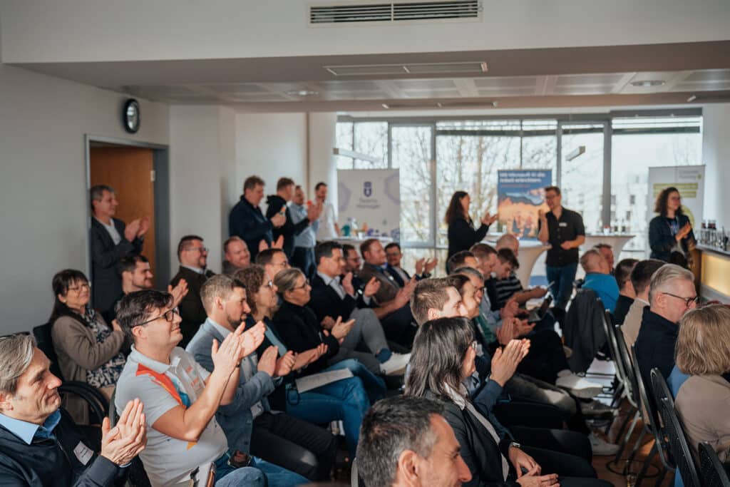 Eine Gruppe von Personen sitzt und steht in einem hellen Konferenzraum, hört aufmerksam zu und applaudiert einer Business Intelligence-Präsentation. Einige halten Notebooks in der Hand, und im Hintergrund sind in der Nähe großer Fenster Banner zu sehen.