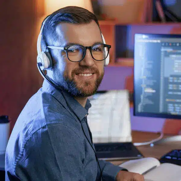 Ein Mann mit Brille und Headset lächelt, während er in einem schwach beleuchteten Büro an einem Schreibtisch mit aufgeschlagenen Notebooks und einem Computermonitor sitzt, auf dem ein Microsoft 365-Code angezeigt wird.