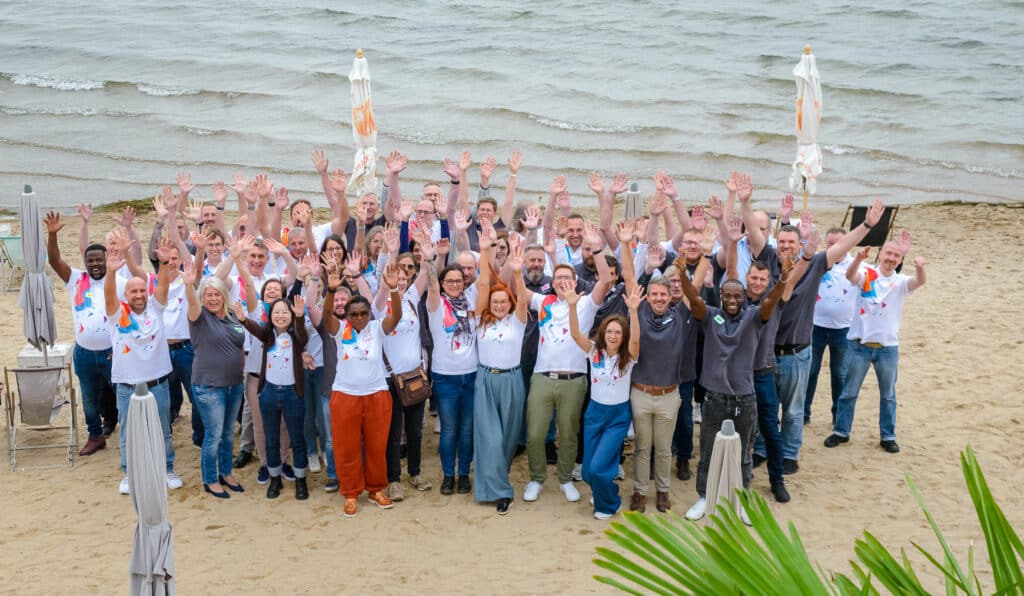 Eine große Gruppe von Menschen steht an einem Sandstrand in der Nähe des Wassers, lächelt und hebt feierlich die Hände. Die meisten tragen passende weiße Hemden mit bunten Motiven und feiern ihren Erfolg mit Microsoft Power BI. Um sie herum sind geschlossene Regenschirme zu sehen.