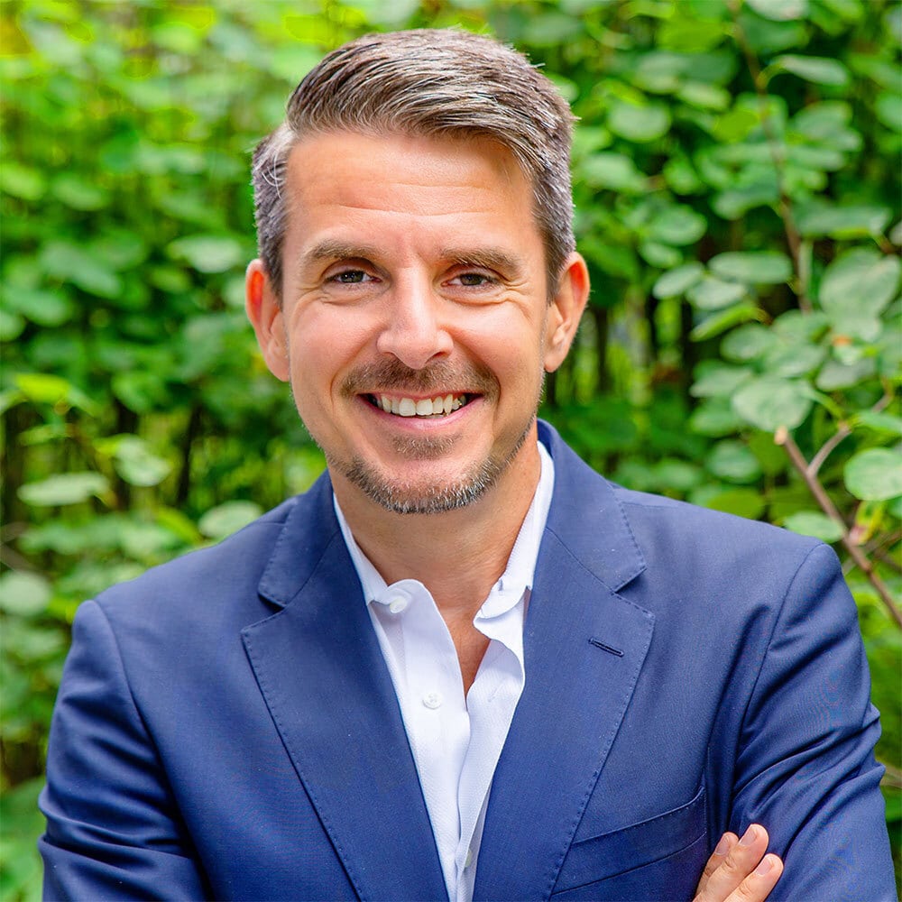 Ein lächelnder Mann mit kurzen, gepflegten Haaren und leichten Bartstoppeln trägt einen marineblauen Blazer und ein weißes Hemd und steht im Freien vor üppig grünem Laub.