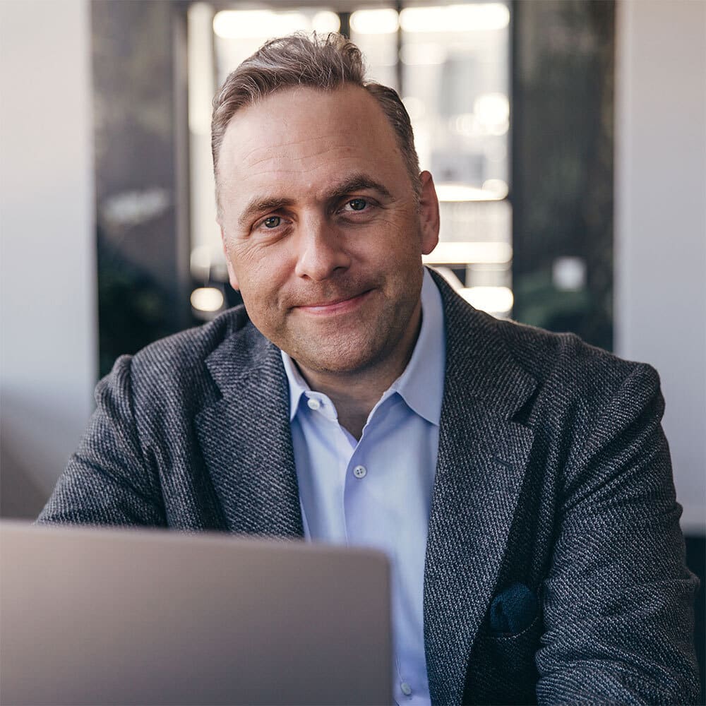 Ein Mann mit kurzen Haaren in einem blauen Hemd und einem strukturierten Blazer sitzt an einem Schreibtisch und lächelt leicht in die Kamera. Vor ihm steht ein Laptop und ein heller, moderner Bürohintergrund ist zu sehen.