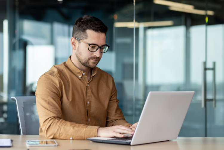 Ein Mann mit Brille und braunem Hemd sitzt an einem Schreibtisch und tippt auf einem Laptop in einem modernen Büro mit Glaswänden.