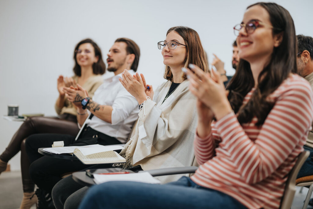 Eine Gruppe von Menschen, die in einem Klassenzimmer sitzen, lächeln und applaudieren. Einige haben Notizbücher und Stifte dabei, was darauf hindeutet, dass sie an einer Vorlesung oder einem Seminar teilnehmen. Die Atmosphäre scheint positiv und engagiert zu sein.