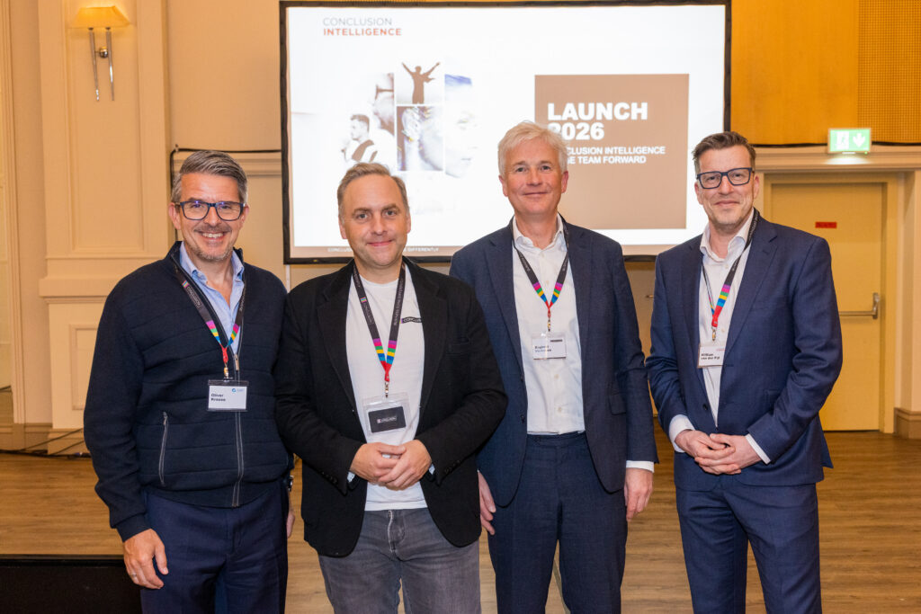 Vier Männer in Geschäftskleidung stehen lächelnd vor einem Präsentationsbildschirm, auf dem "Launch 2026 Conclusion Intelligence" in einem Konferenzraum zu sehen ist.