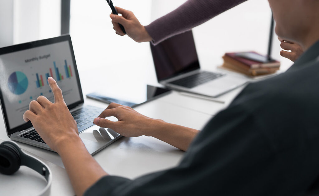 Zwei Personen arbeiten an einem Schreibtisch mit Laptops. Eine Person zeigt auf einen Laptop-Bildschirm, auf dem bunte Diagramme und Grafiken angezeigt werden, während die andere Person mit einem Stift gestikuliert. Auf dem Schreibtisch befinden sich auch Kopfhörer und Bücher.