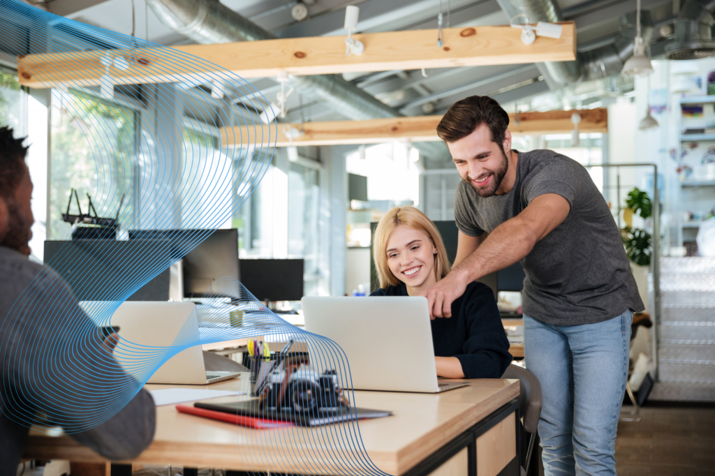 Ein Mann steht neben einer lächelnden Frau, die an einem Schreibtisch sitzt und auf den Bildschirm ihres Laptops zeigt – in einem hellen, modernen Büro. Eine weitere Person sitzt ihnen gegenüber und arbeitet am Computer.